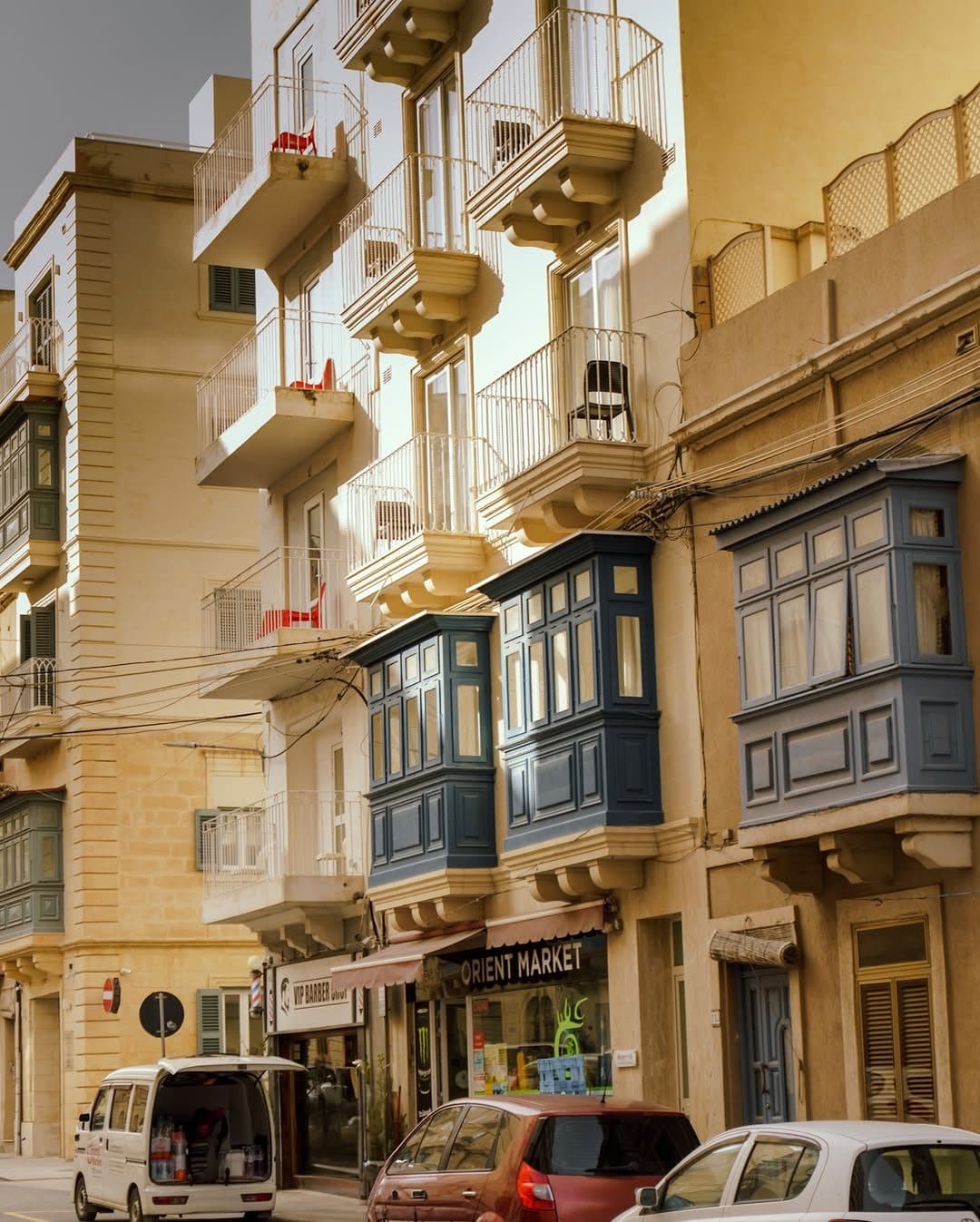 Street photo by Chiemeka Madufor: sunlit limestone townhouses with enclosed wooden balconies in blue and green, cars parked below