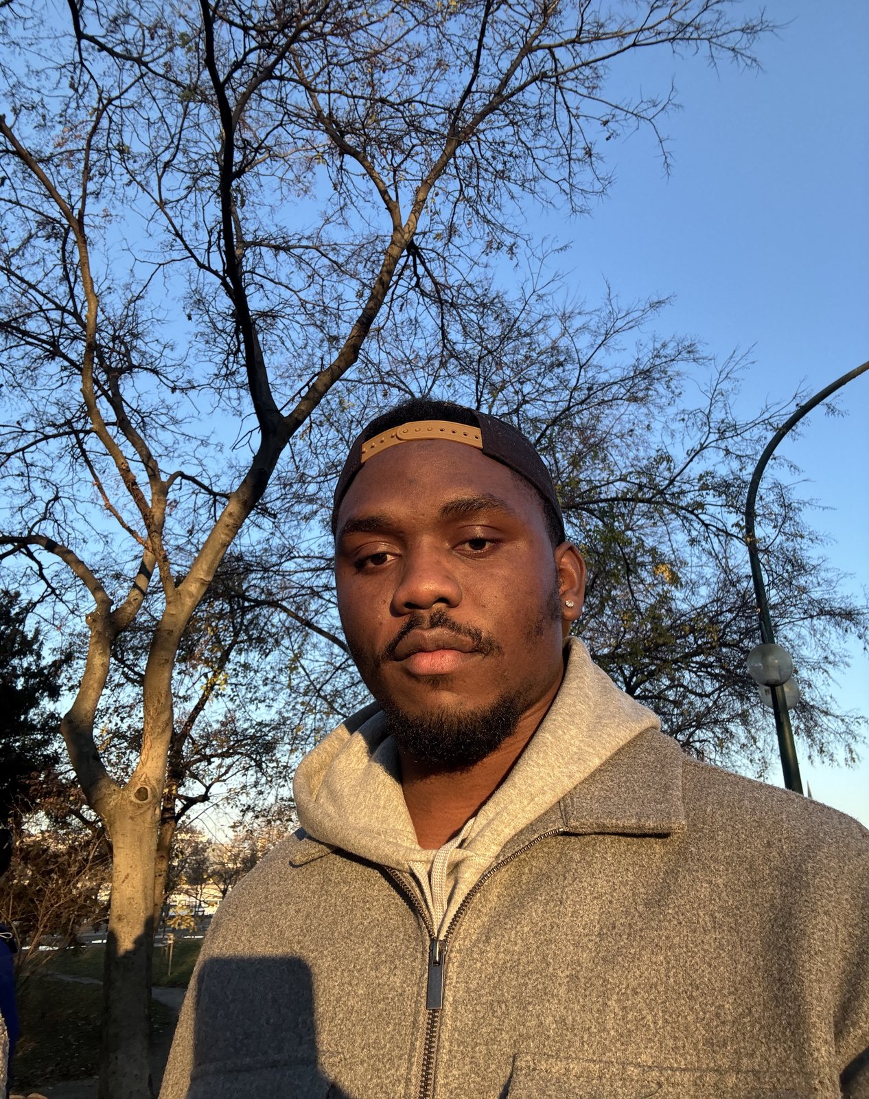Chiemeka Madufor in a cap and grey jacket under a bare tree in the evening sun